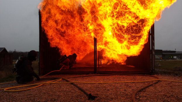 Feuerwehrleute löschen ein Gebäudebrand mit einem Schlauch auf dem Boden, Schuppen und Bäume sind im Hintergrund unter einem klaren blauen Himmel sichtbar.