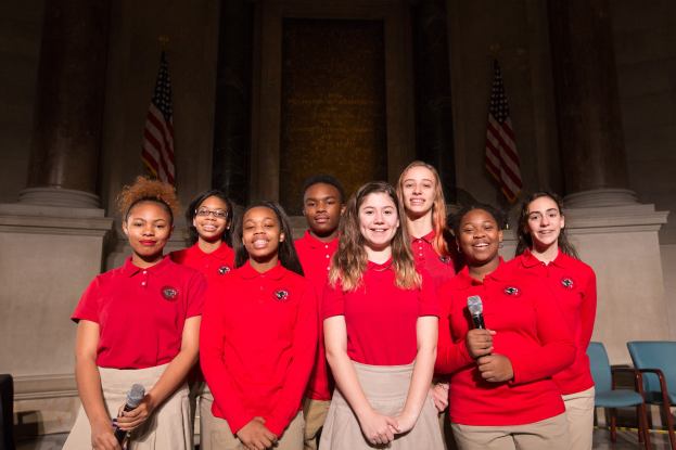 Eine Gruppe junger Frauen in roten Hemden, die strahlend Mikrofone halten und vor Stühlen, Säulen und Flaggen stehen, wahrscheinlich Teil eines nationalen Jugendchors.