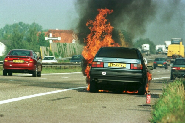 Ein Auto in Flammen auf der Seite der Straße, umgeben von anderen Fahrzeugen, mit Bäumen, Gebäuden und einem klaren blauen Himmel im Hintergrund und Gras und einem Feuerlöscher auf der rechten Seite.