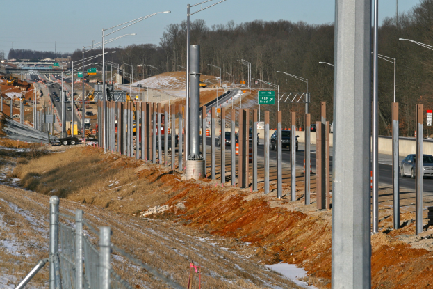 Baustelle mit mehreren Fahrzeugen, Pfählen, beleuchteten Schildern, einem Zaun, schneebedecktem Gras, Bäumen und einem Himmel im Hintergrund.