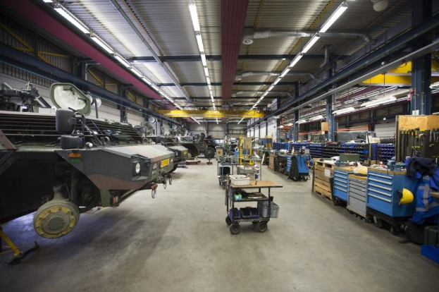 Großer Lagerinnenraum mit Maschinen, Fahrzeugen auf der linken Seite, Transportwagen und Holzboxen auf der rechten Seite sowie Deckenbeleuchtung.