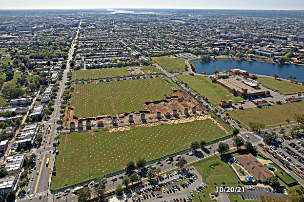 Luftaufnahme einer Stadt mit Gebäuden, Bäumen, Gras, Straßen mit Fahrzeugen, Wasser und bewölktem Himmel, die ein großes zentrales Feld als Parkplatz zeigt.
