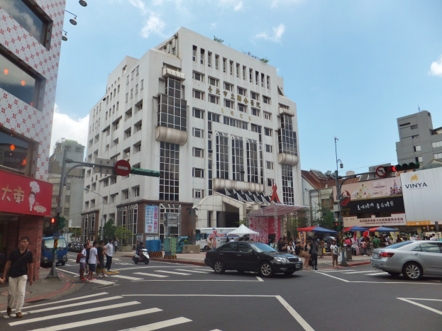 Eine belebte Straße mit Fussg"angern, die vor einem hohen Geb"ude die Straße "uberqueren, Fahrzeuge auf der Straße und Verkehrszeichen, mit Gehwegen, Geb"uden, Schildern, Schirmen, B"umen und einem bew"olkten Himmel im Hintergrund.