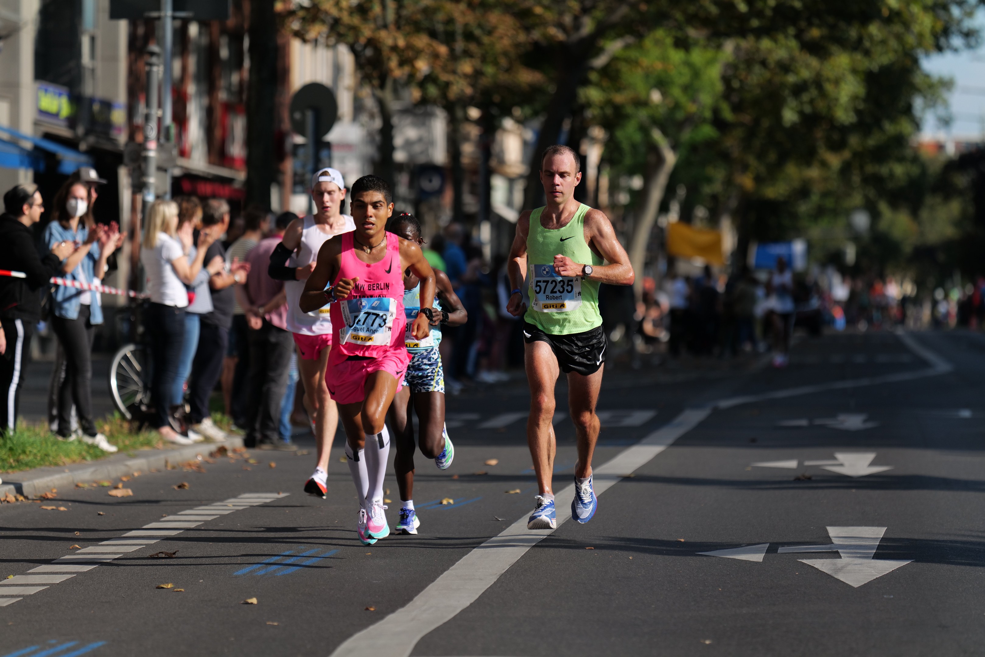 Gruppe von Menschen beim Marathon auf einer Stadtstraße mit Zuschauern, Bäumen, Gebäuden und einem Fahrrad im Hintergrund.
