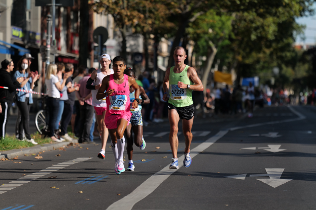 Gruppe von Menschen beim Marathon auf einer Stadtstraße mit Zuschauern, Bäumen, Gebäuden und einem Fahrrad im Hintergrund.