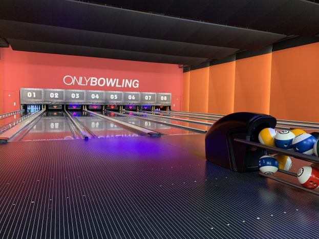 Eine Bowlingbahn mit Bowlingkugeln auf der rechten Seite, Bowlingpins in der Mitte und einer Wand mit Text im Hintergrund, unter einer sichtbaren Decke.