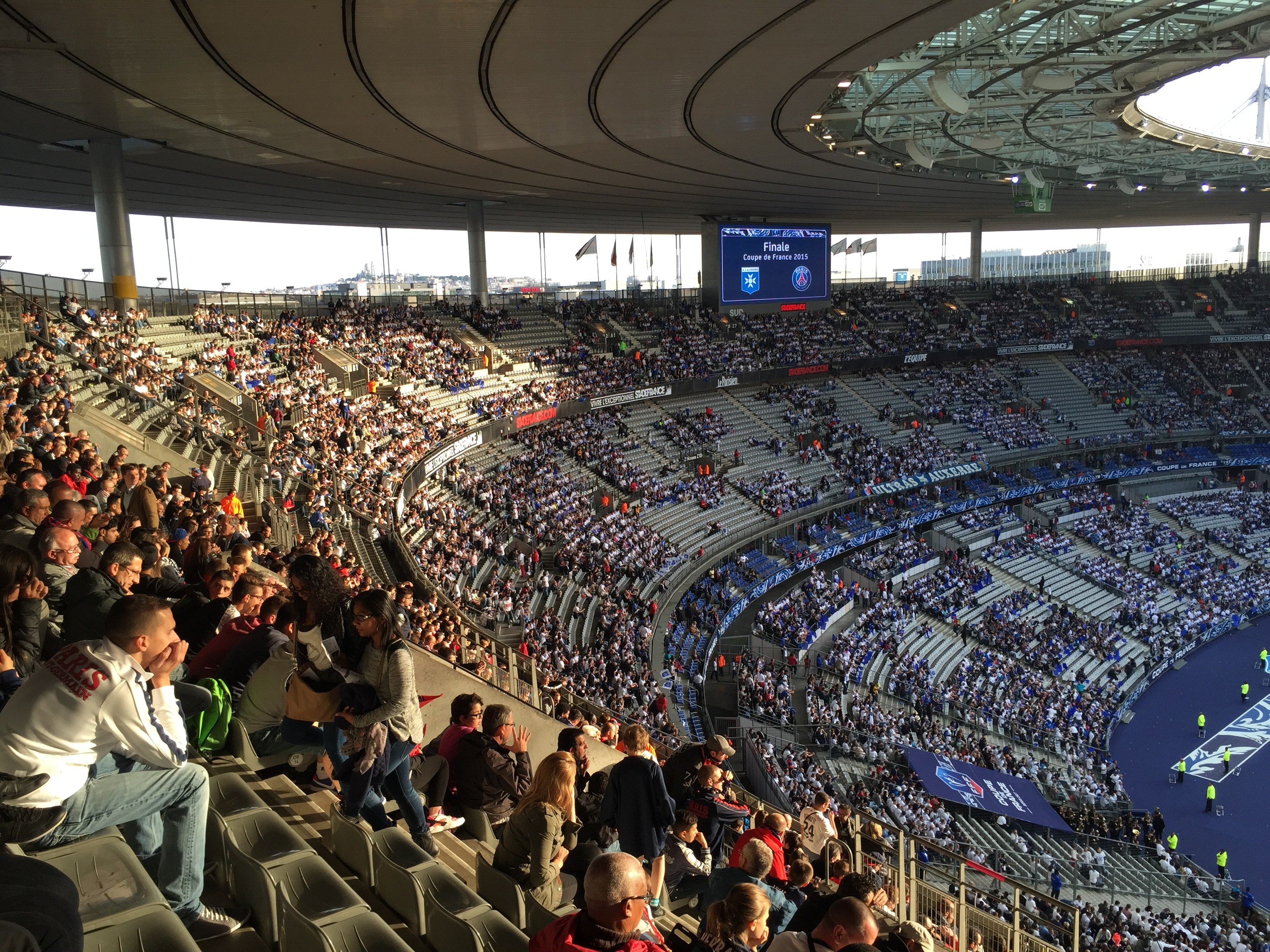 Große Menschenmenge in einem Stadion bei einem Fußballspiel, mit einer Bühne rechts, Fahnen, Stangen, einem Bildschirm und der Allianz Arena in München, Deutschland im Hintergrund.