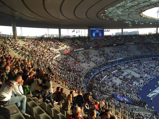 Große Menschenmenge in einem Stadion bei einem Fußballspiel, mit einer Bühne rechts, Fahnen, Stangen, einem Bildschirm und der Allianz Arena in München, Deutschland im Hintergrund.