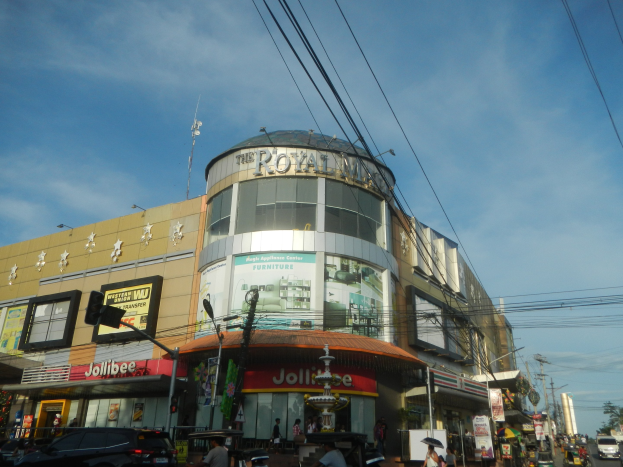 Eine belebte Stadtstraße mit fahrenden Fahrzeugen, Fußgängern auf dem Gehweg und dem Royal Mall-Einkaufszentrum im Hintergrund, umgeben von Bäumen, Versorgungsmasten und Leitungen unter dem Himmel.