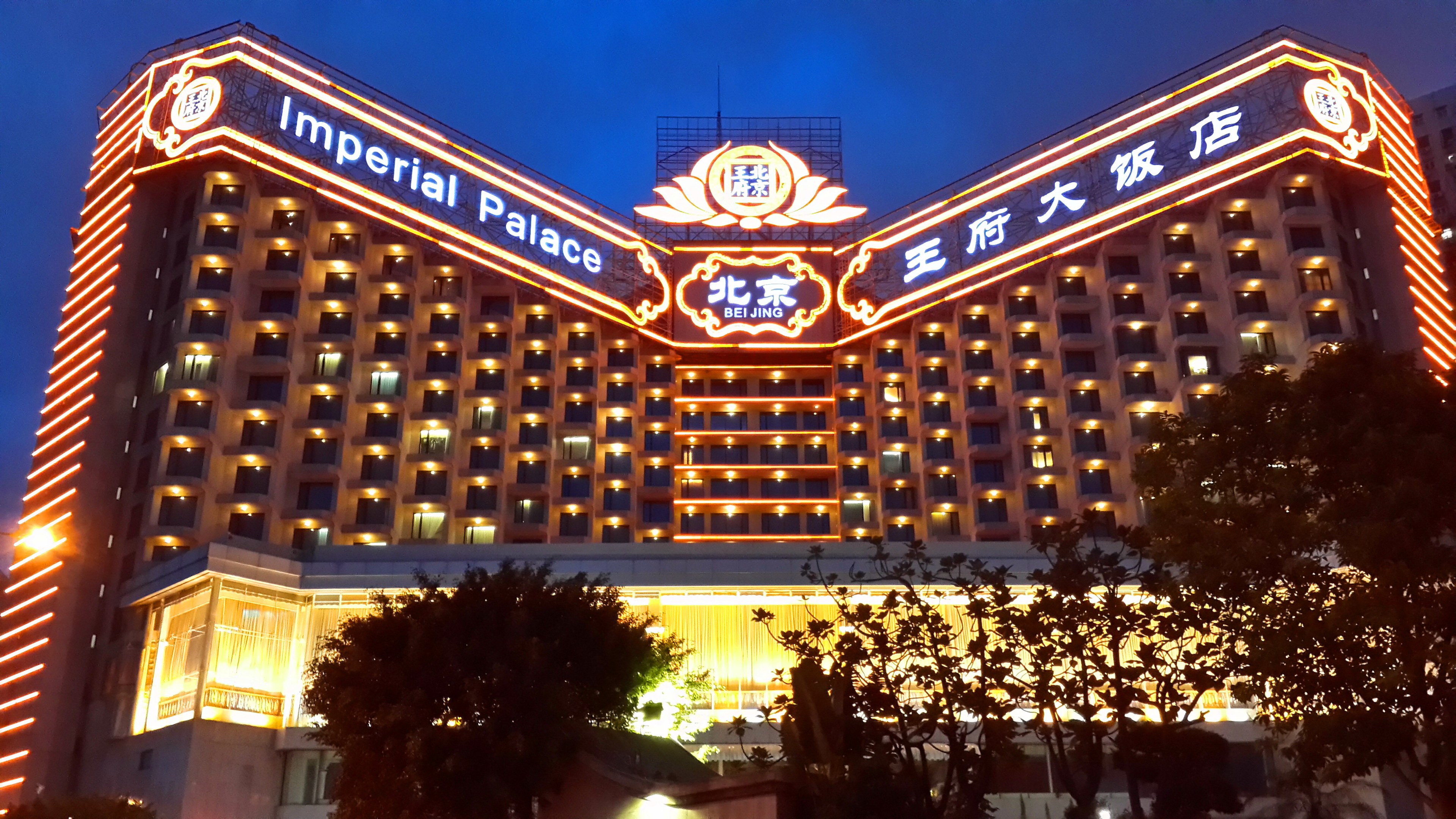 Das Hotel Imperial in Peking, China, ein großes mehrstöckiges Gebäude mit zahlreichen Fenstern und beleuchteter Beschilderung, vor einem klaren blauen Himmel mit Bäumen im Vordergrund.