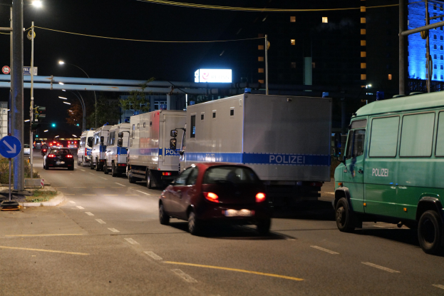 Eine Reihe von Polizeifahrzeugen steht nachts am Straßenrand, mit Gebäuden, einer Brücke und Bäumen im Hintergrund.