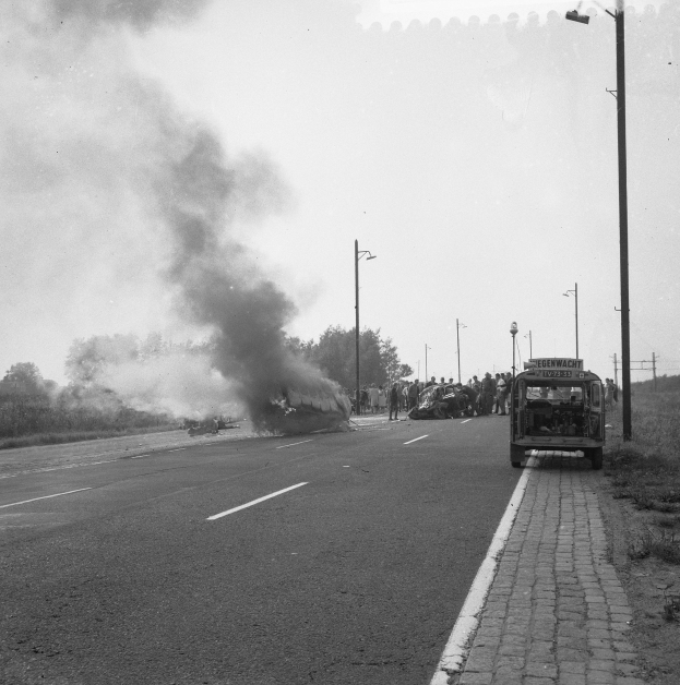 Schwarz-weiß-Foto eines brennenden Busses auf einer Straße mit einigen Menschen in der Nähe, Polen, Bäume und Himmel im Hintergrund.