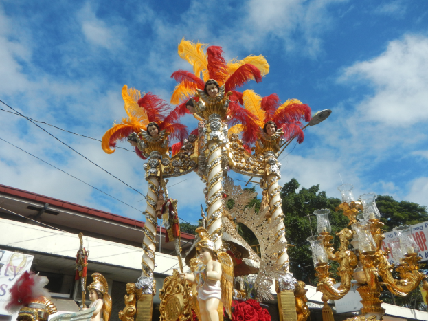 Ein Karnevalswagen, geschmückt mit goldenen und roten Federn, trägt Menschen und ist in einem Umzug mit einem Gebäude, Bäumen, Drähten und einer Fahne in der Nähe zu sehen, unter einem sichtbaren Himmel.