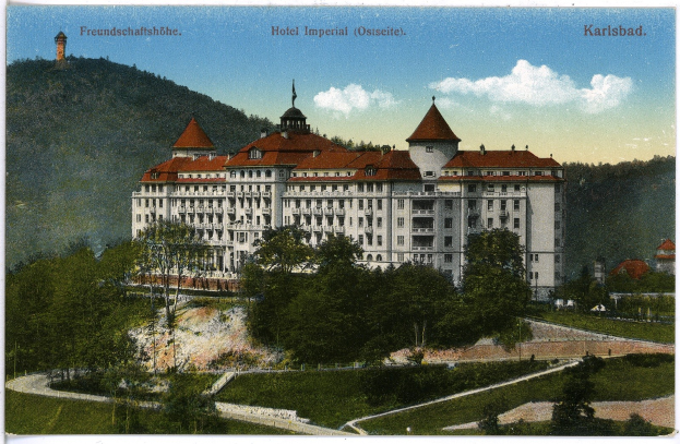 Eine alte Postkarte des Hotel Imperial in Karlsbad, Deutschland, zeigt ein großes Gebäude mit Fenstern, umgeben von Bäumen, Pflanzen, Gras, einem Weg, einer Gruppe von Bäumen, Hügeln und einem bewölkten Himmel, mit Text oben.