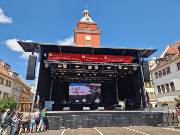 Gruppe von Menschen auf einer Bühne vor einem Uhrenturm auf dem Kreissparkasse Gotha Musikfestival, mit Gebäuden, Bäumen und einem bewölkten Himmel im Hintergrund.