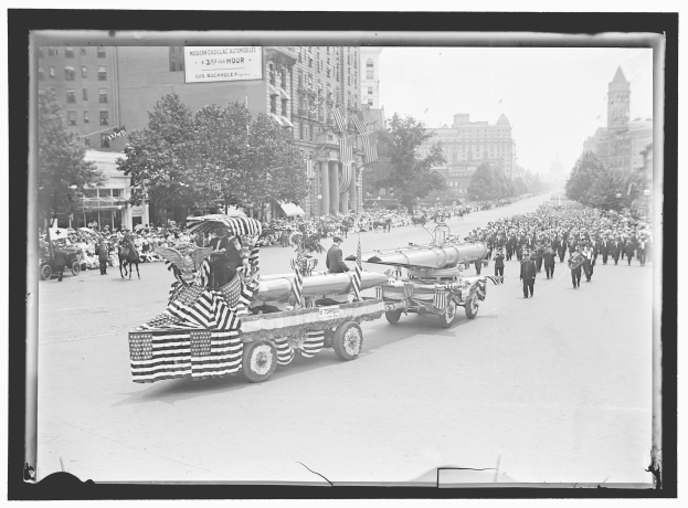 Schwarzes und weißes Foto eines Umzugs mit einem Feuerwehrauto in der Mitte, umgeben von Menschen mit Fahnen, sowie Bäumen, Gebäuden und einer Tafel im Hintergrund.