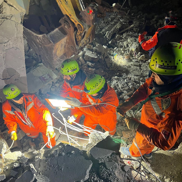 Eine Gruppe von Menschen in orangefarbenen Uniformen und grünen Helmen bei der Arbeit an Schutt von einem eingestürzten Gebäude, im Hintergrund ein Kran, mit Rettungsgeräten und -materialien.