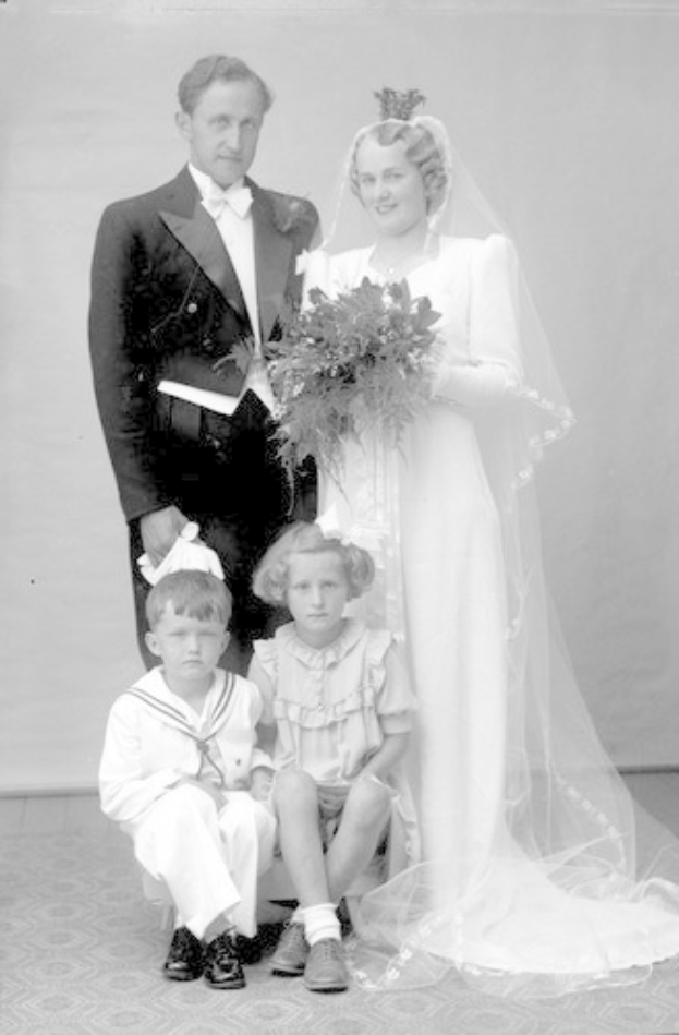 Schwarz-weiß-Foto einer Braut in einem weißen Kleid mit einem Strauß, Bräutigam, der neben ihr lächelt, und zwei Kindern, die vor ihnen auf dem Boden sitzen.