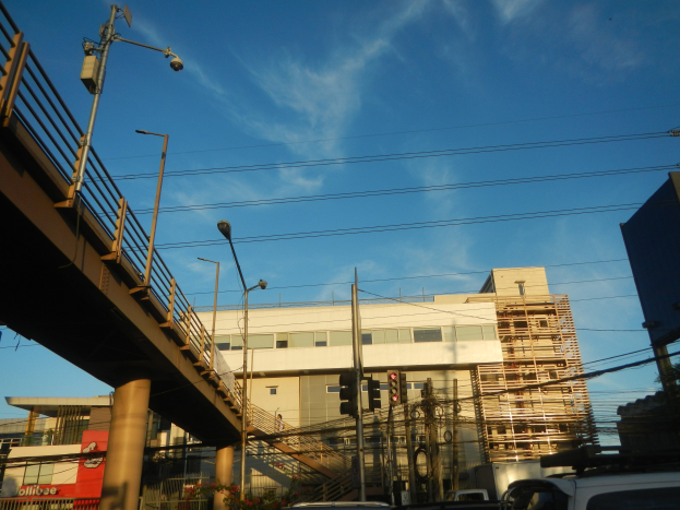 Stadtstraße mit parkenden Autos, eine Brücke mit Geländern, Laternen, Strommasten mit Kabeln, Ampeln, Gebäude mit Fenstern und ein bewölkter Himmel im Hintergrund.
