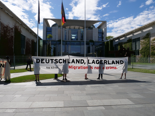 Gruppe von Menschen mit einem Transparent "Deutschland, Lagerland Migration ist kein Verbrechen" vor einem Gebäude mit Fenstern, umgeben von Gras, einem Metallzaun, Pflanzen, Bäumen und Fahnenstangen.